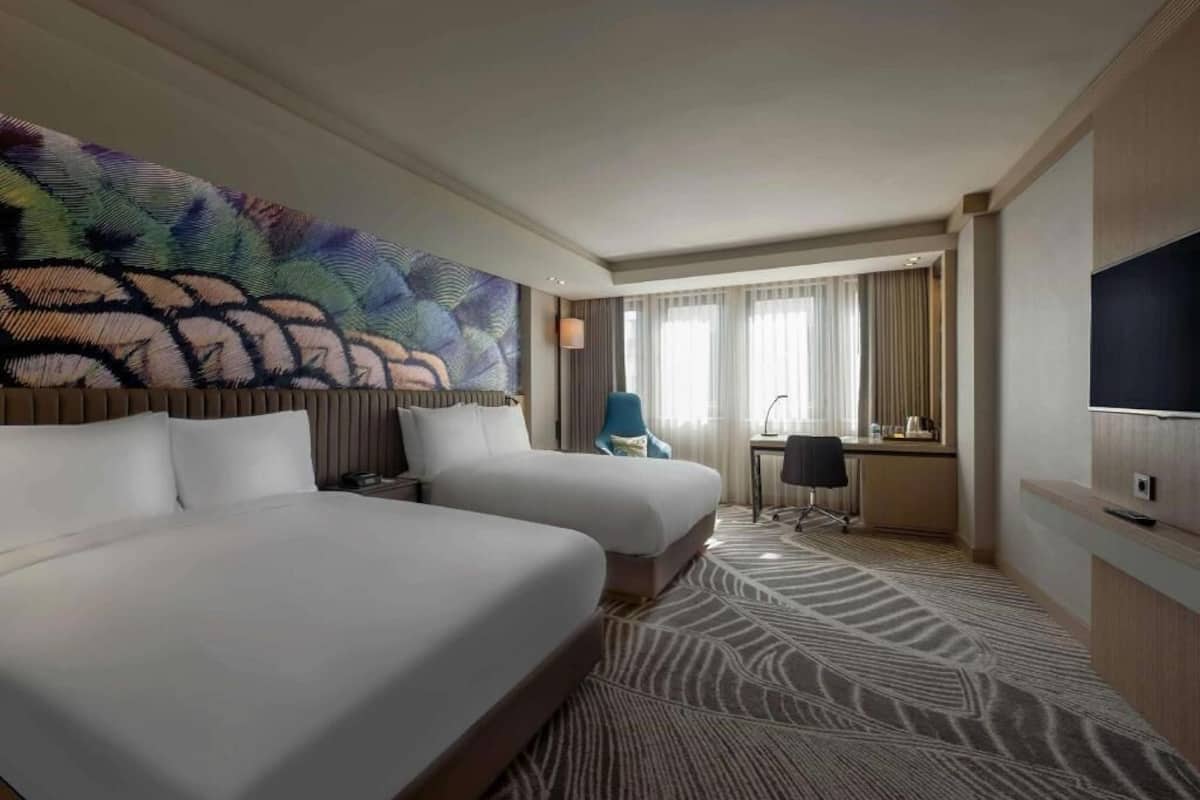 A hotel room with two beds, a desk, a chair, a television, and a large abstract wall art piece.
