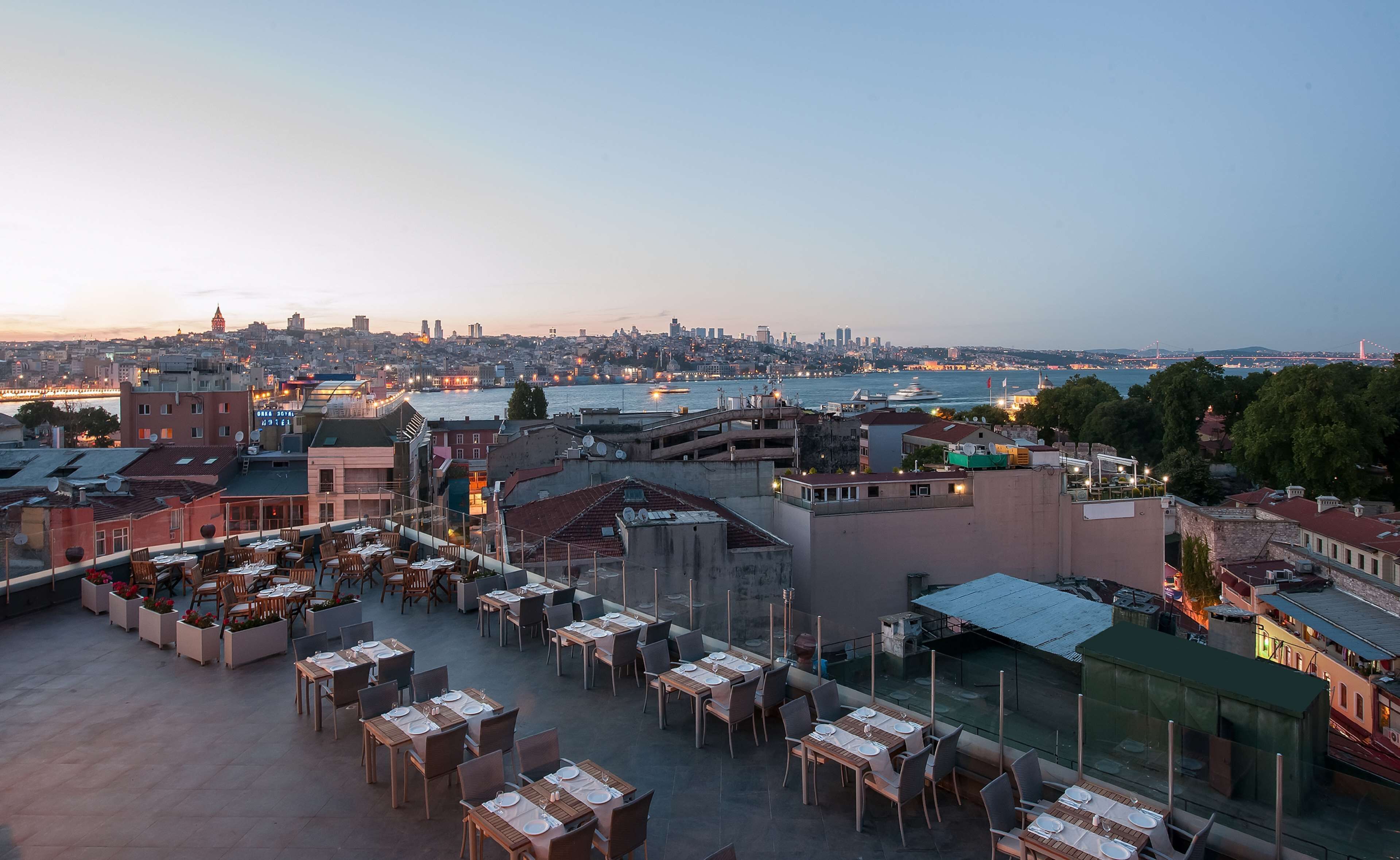 Un área de comedor al aire libre con varias mesas y sillas, con vista a un paisaje urbano y una masa de agua.