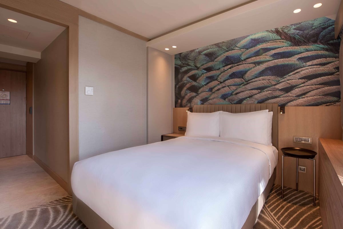 A hotel room with a large bed, a headboard with a leaf-patterned mural, a wooden nightstand, and a carpeted floor.