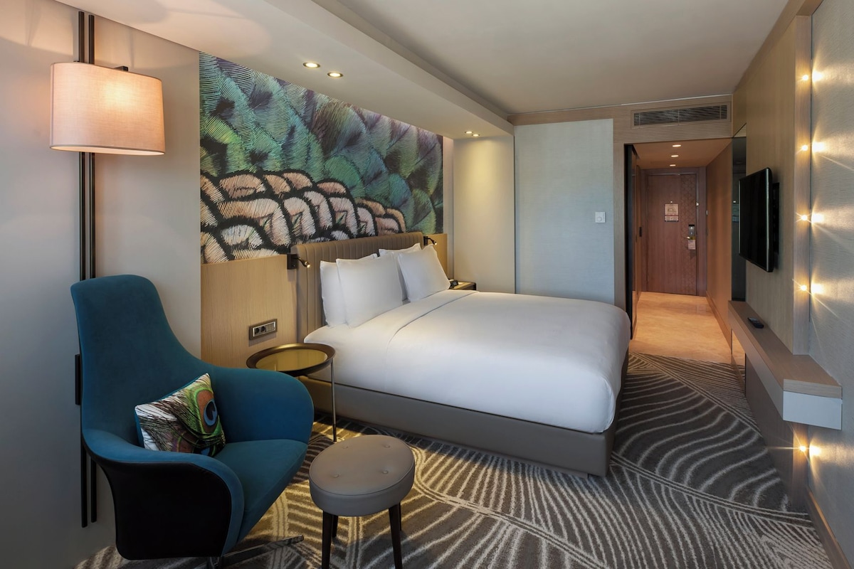 A hotel room with a bed, a seating area with a chair and ottoman, a wall-mounted TV, and a patterned carpet.