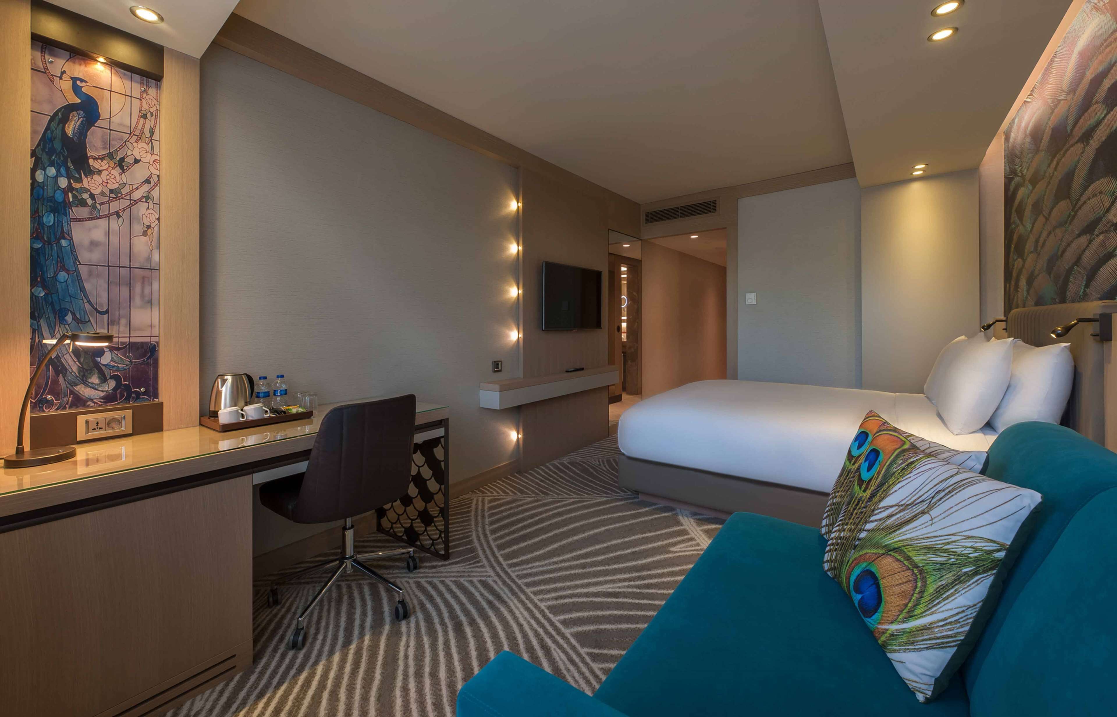 A hotel room with a bed, a desk with a chair, a TV, and a peacock-themed pillow.