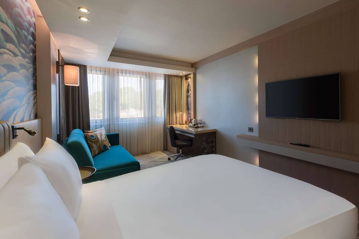 A hotel room with a large bed, a desk with a chair, a television mounted on the wall, and a window with curtains.