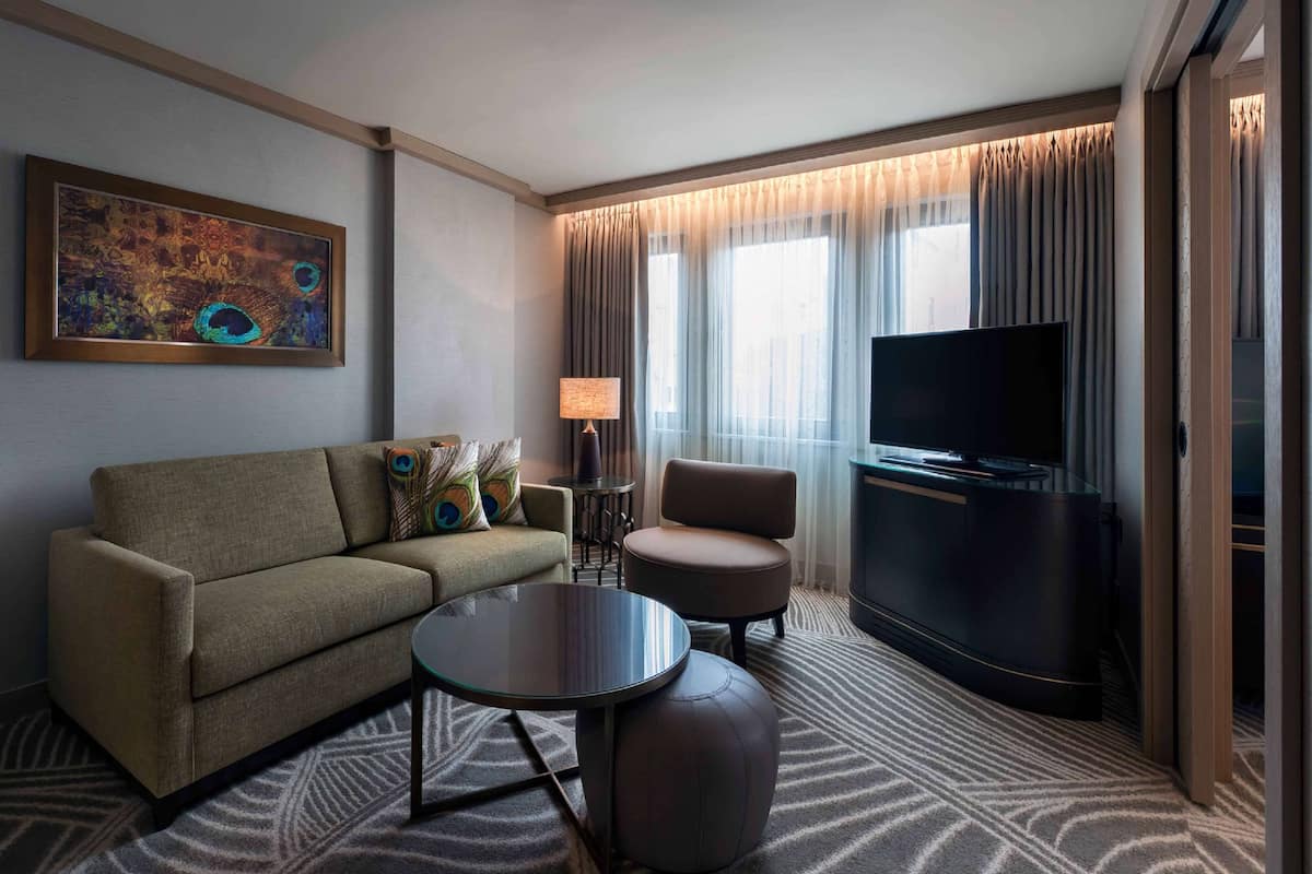 A hotel room with a sofa, armchair, TV, lamp, and artwork on the wall.