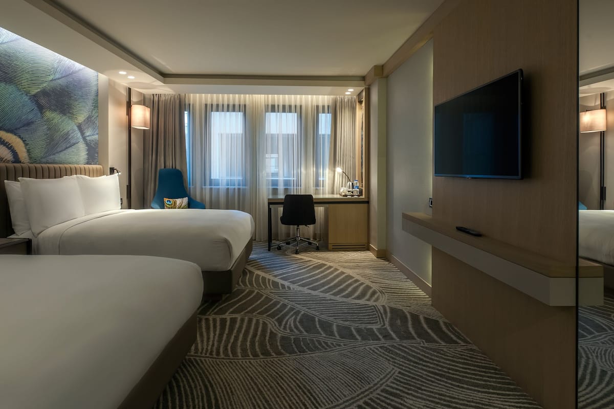 A hotel room with two beds, a desk, a TV, and a large window with curtains.