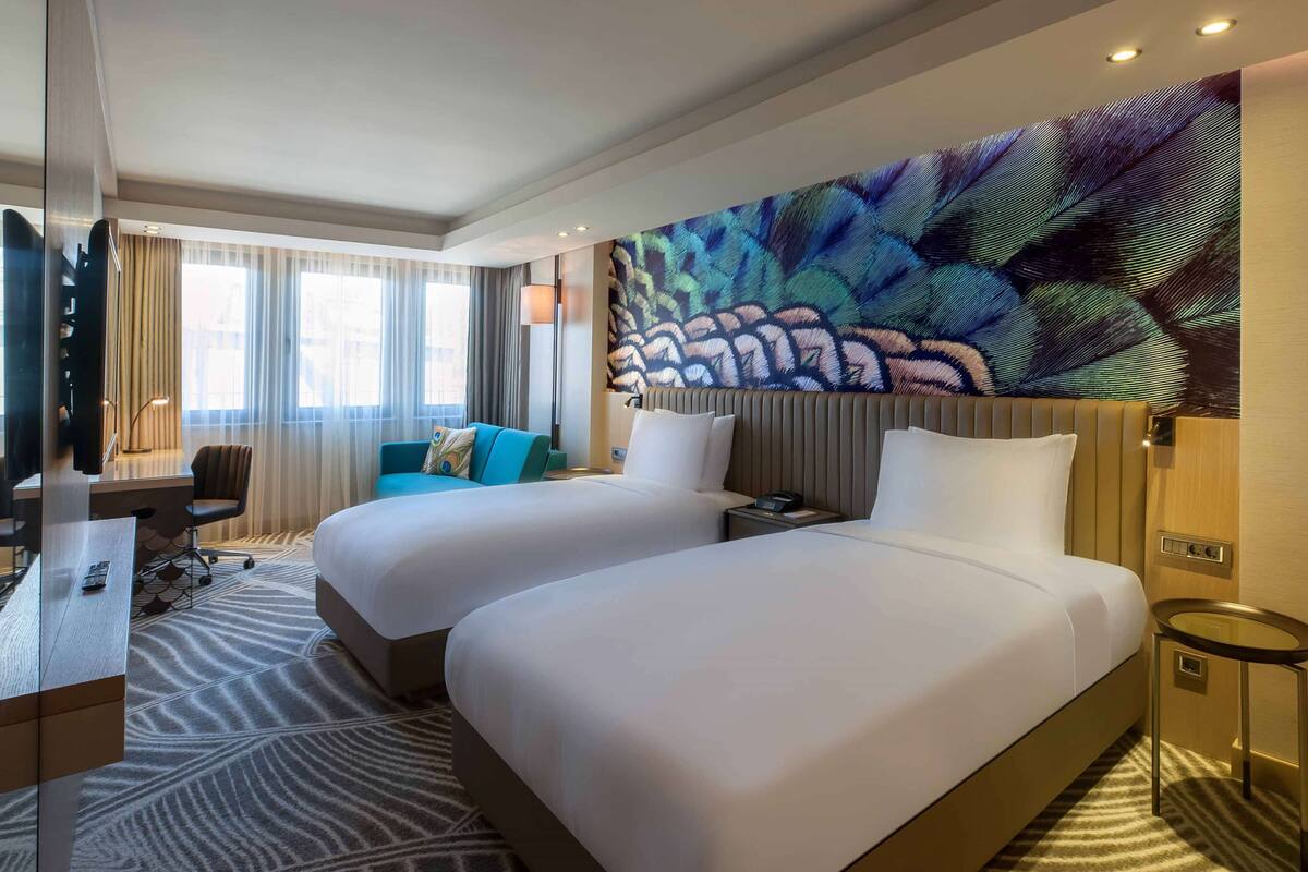 A hotel room with two beds, a desk, a chair, a TV, and a large abstract wall mural.