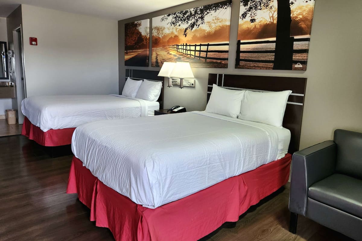 A hotel room with two beds, each with white bedding and red skirts, a nightstand with a lamp, and a wall-mounted artwork depicting a sunset scene.