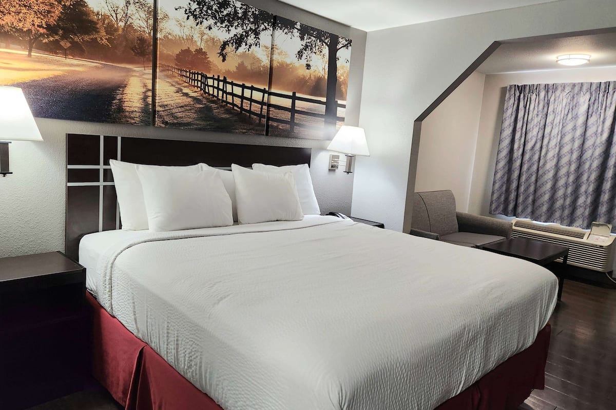 A hotel room with a large bed, a headboard with storage, a desk, a chair, a wall-mounted lamp, and a window with curtains.
