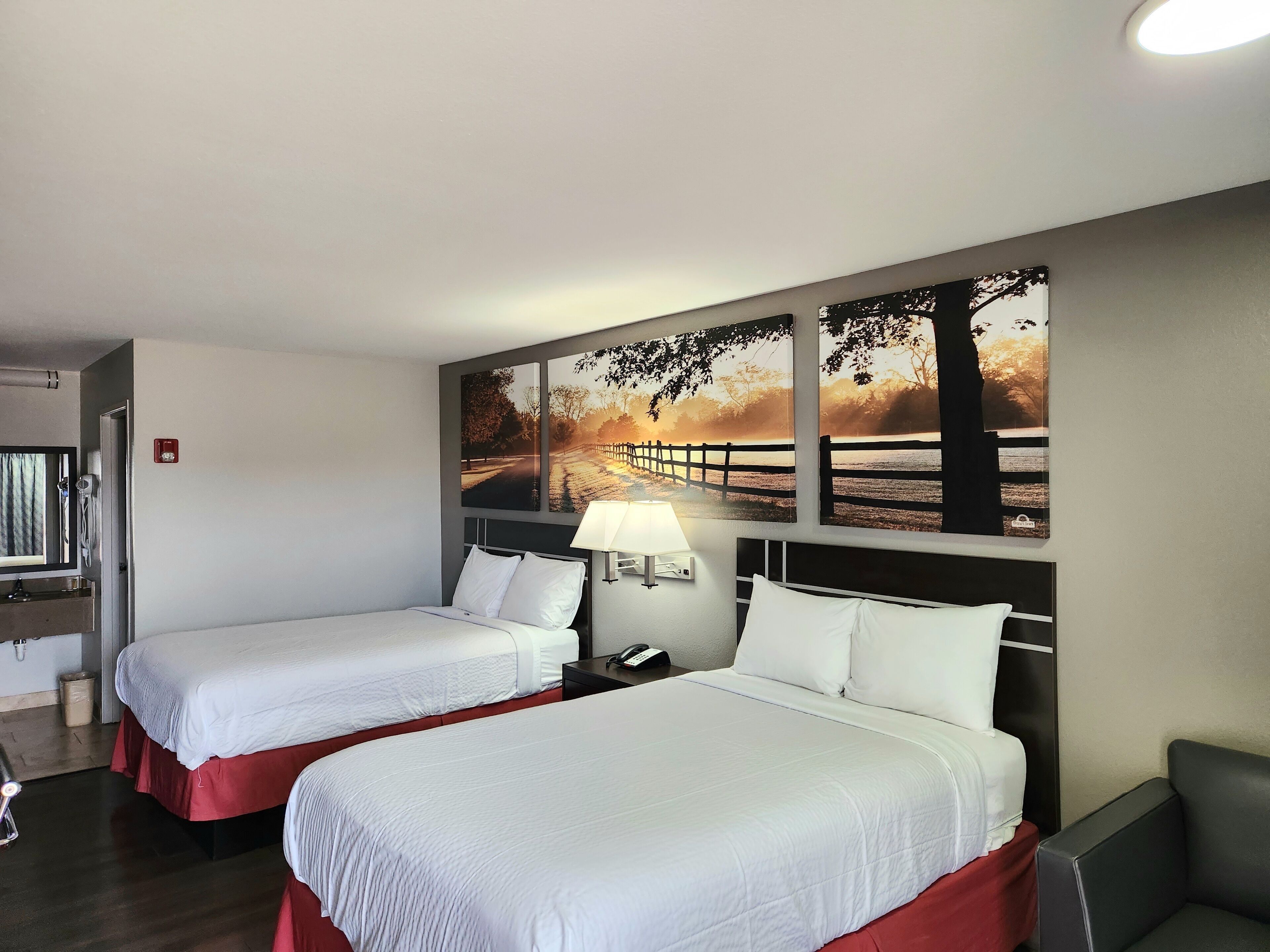 Ein Hotelzimmer mit zwei Betten, einem Nachttisch, einer Lampe und einem Wandbild, das eine Sonnenuntergangsszene zeigt.