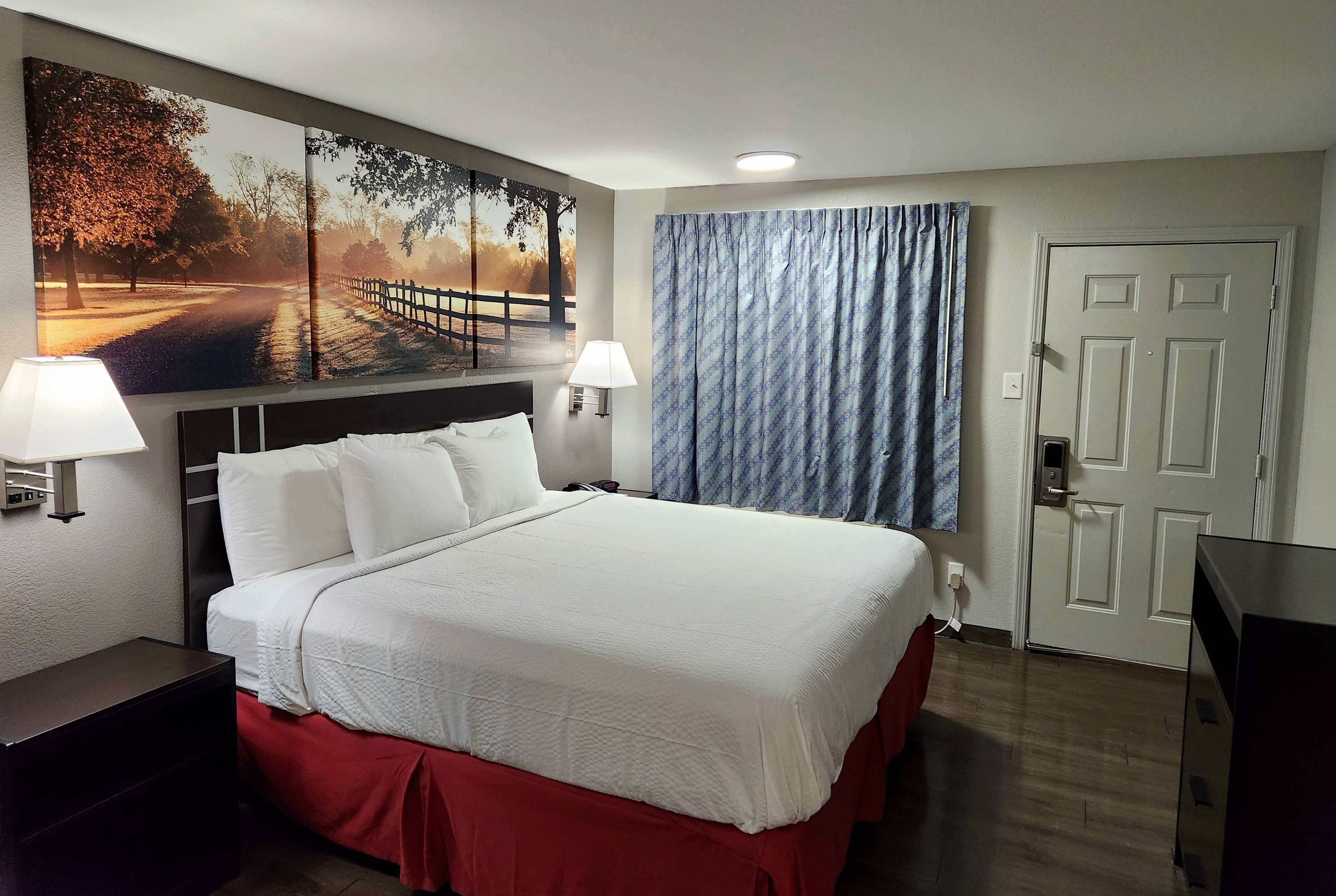 A hotel room with a large bed, two bedside lamps, a nightstand, a door, and a wall-mounted TV.