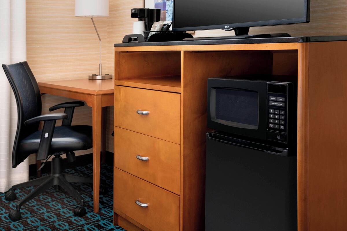 A compact hotel room with a wooden desk, a black office chair, a microwave, and a small refrigerator.