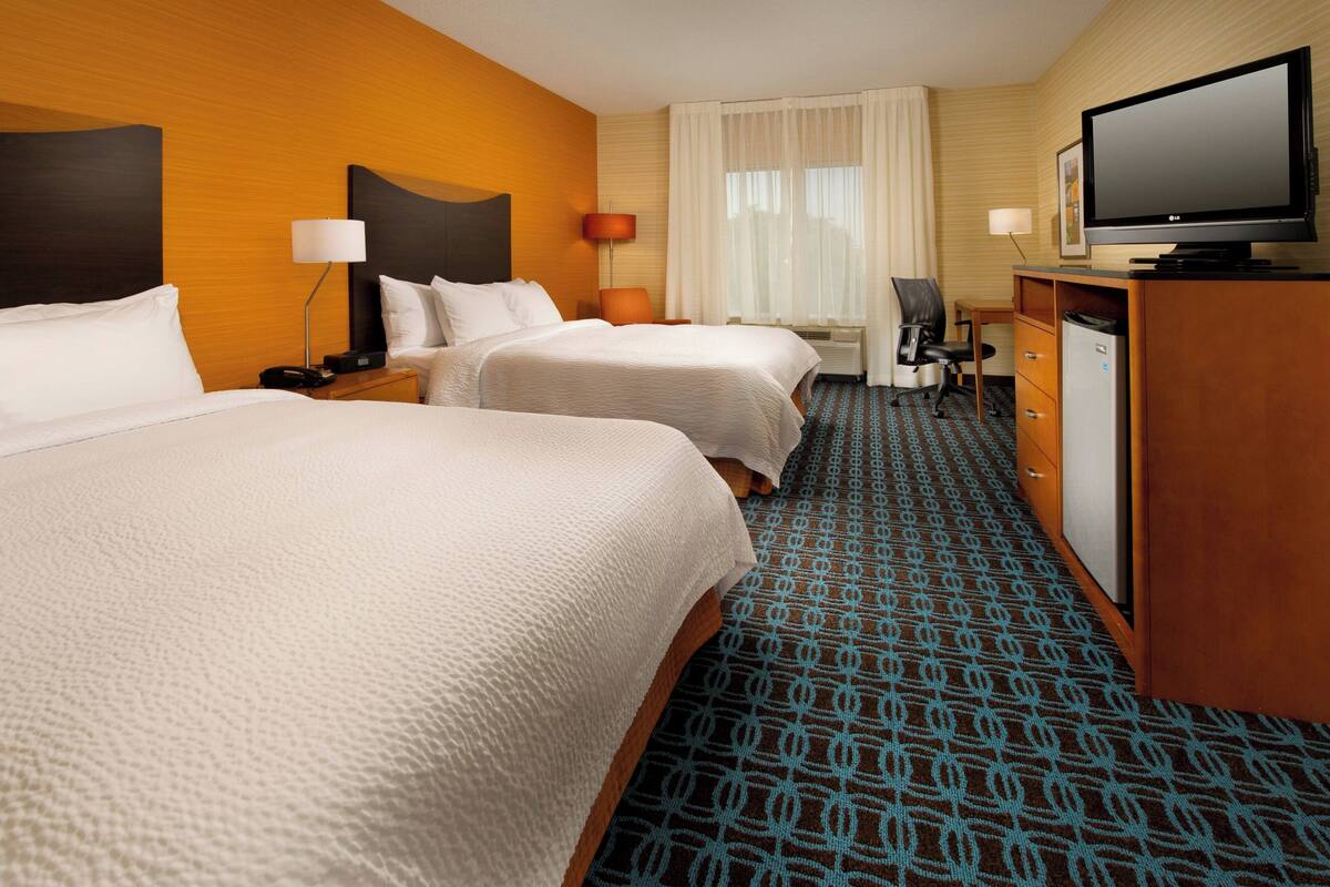 A hotel room with two beds, a desk with a television, a refrigerator, and a patterned carpet.