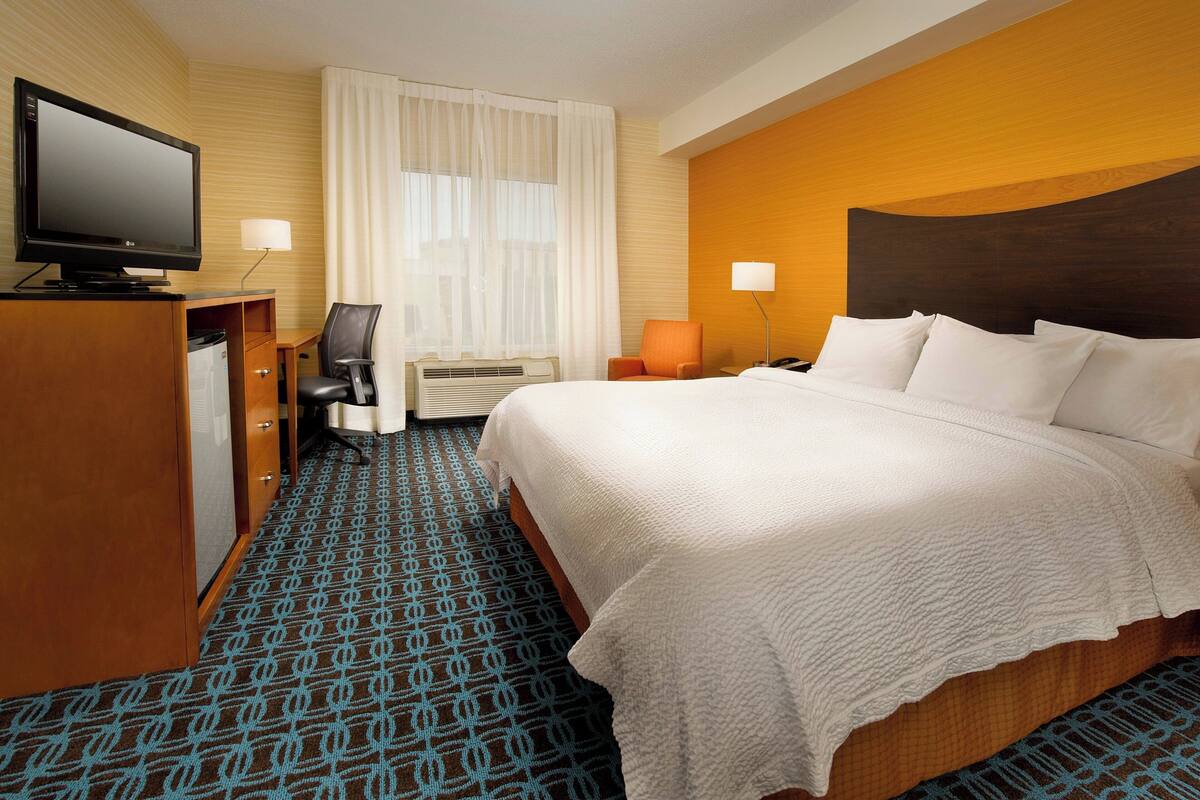 A hotel room with a bed, a desk with a television, a chair, and a lamp.