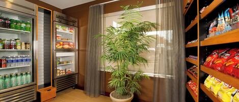 A room with a refrigerator stocked with beverages, a potted plant, and shelves with snacks.