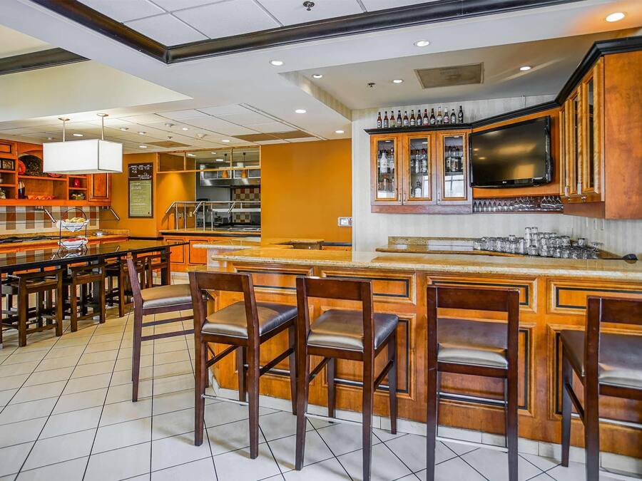 A bar area with wooden bar stools, a television mounted above the bar, and a variety of bottles and glasses on the shelves.