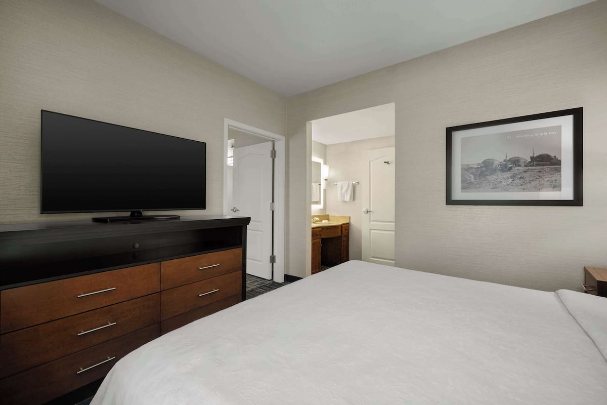 Habitación de hotel con cama, televisor sobre una cómoda de madera, un cuadro en la pared y un baño visible a través de una puerta abierta.