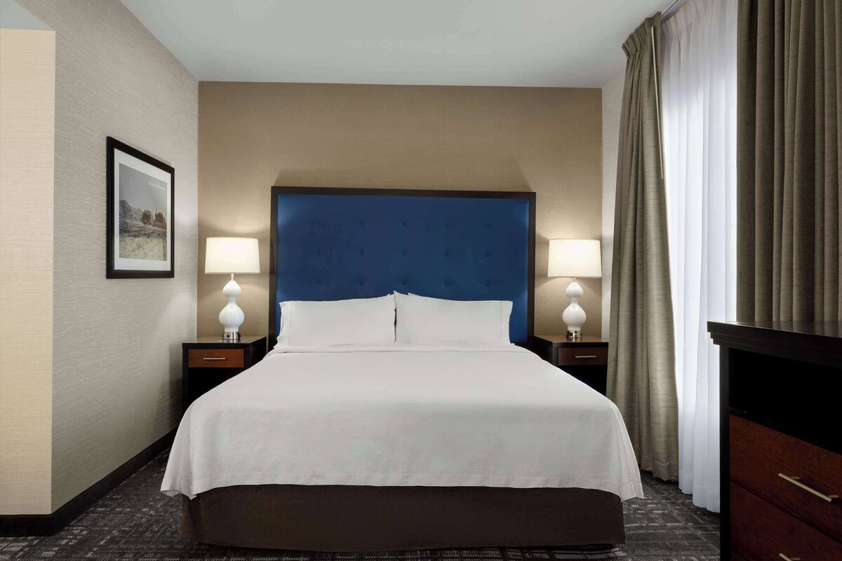Habitación de hotel con una cama grande, dos mesitas de noche con lámparas, una mesita auxiliar, un aparador y un cuadro en la pared.