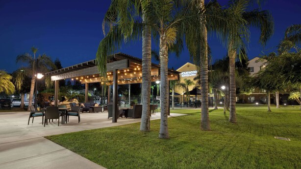 An outdoor patio area with seating and tables, surrounded by palm trees and well-lit by string lights. - Homewood Suites by Hilton Bakersfield (Bakersfield)