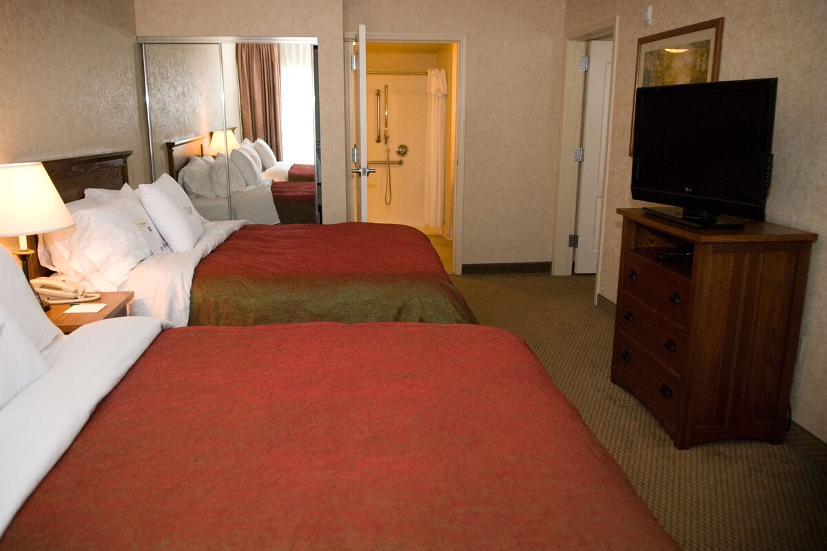 Habitación de hotel con dos camas, televisión, espejo, puerta hacia el baño y mesita de noche con lámpara.