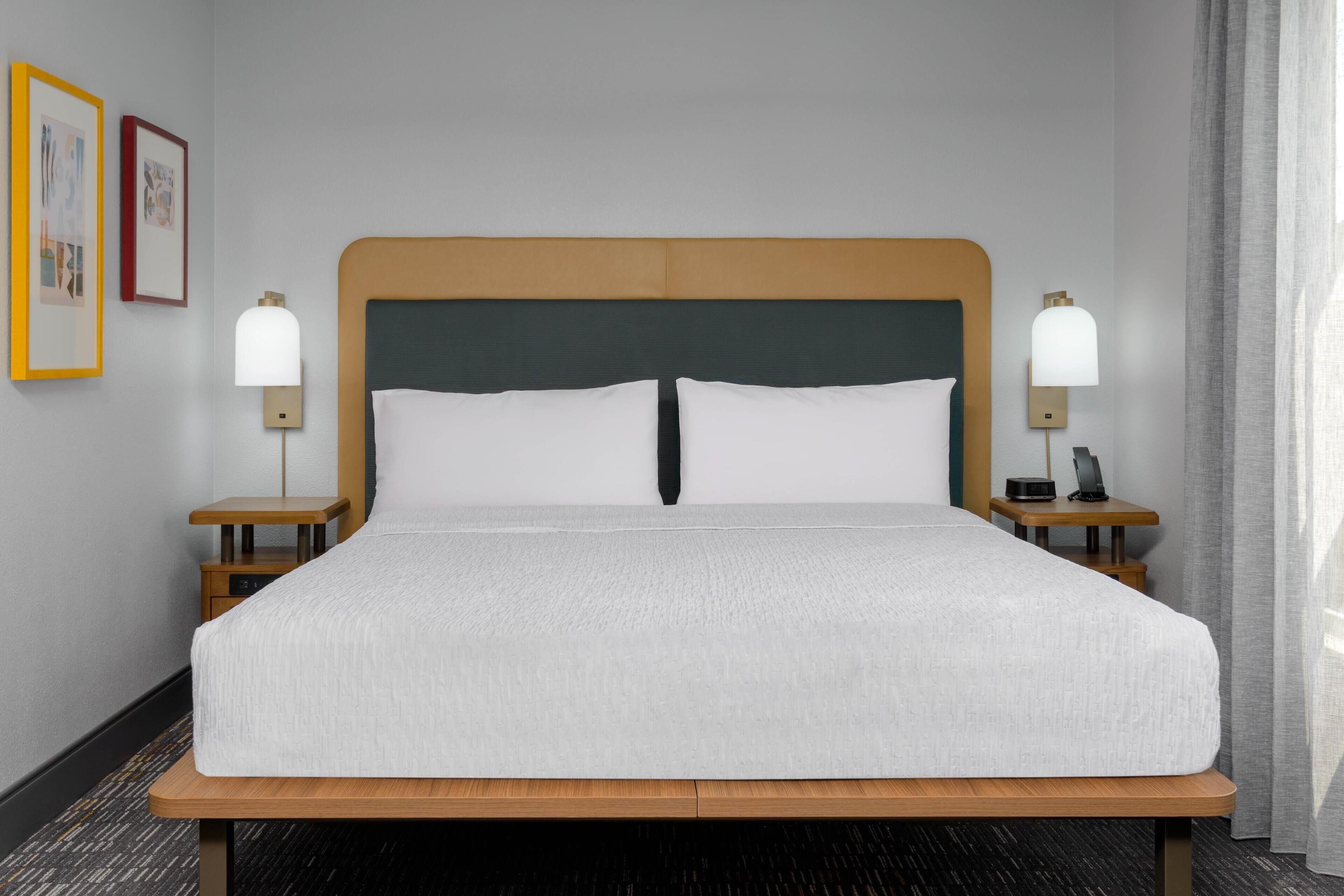 Een net opgemaakt bed met een houten hoofdeinde, twee nachtkastjes met lampen, en ingelijste foto's aan de muur.