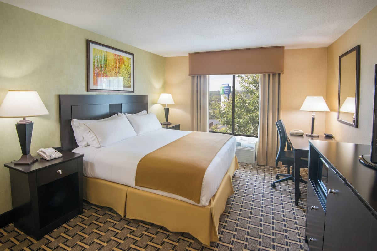 A hotel room with a bed, two bedside tables with lamps, a desk with a chair, and a large window with curtains.