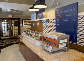 A breakfast station with a flat-top griddle, a sign indicating hot food, and a wall with directional arrows and text. - Holiday Inn Express & Suites Sharon-Hermitage by IHG (West Middlesex)