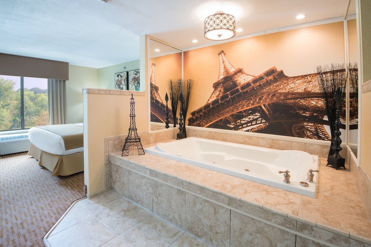 A modern bathroom with a large bathtub, a mirror with an Eiffel Tower mural, and a view of trees through a window.