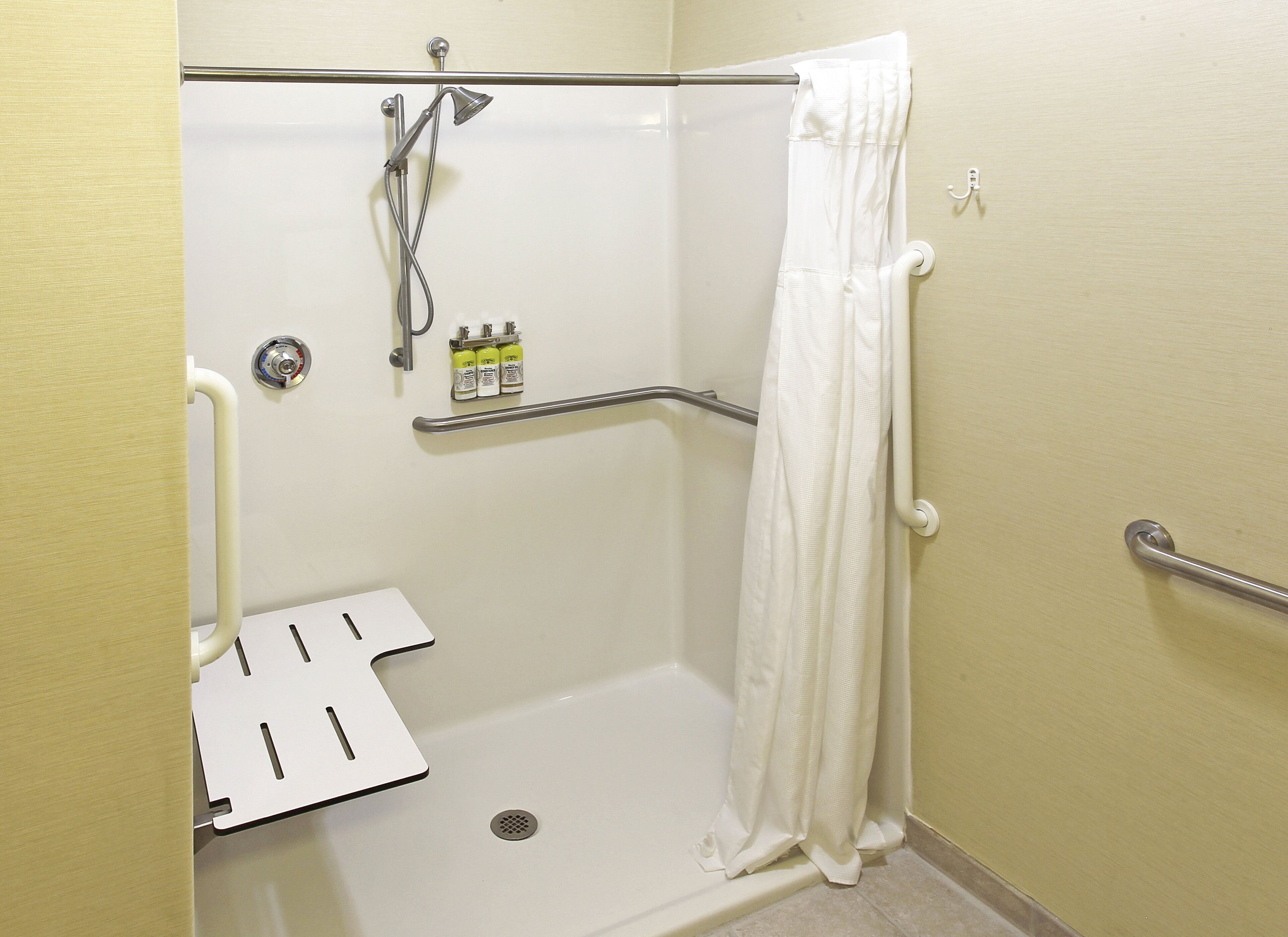 Une douche accessible aux personnes en fauteuil roulant, équipée d’une barre d’appui, d’un rideau de douche blanc et d’une étagère contenant des articles de toilette.