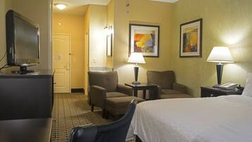A hotel room with a bed, two armchairs, a desk with a television, a lamp, and a painting on the wall.
