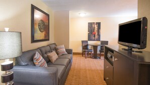 A hotel room with a grey sofa, a TV, a lamp, a dining table with chairs, and a carpeted floor.