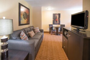 A hotel room with a grey sofa, a TV, a lamp, a dining table with chairs, and a carpeted floor. - Holiday Inn Express & Suites Sharon-Hermitage by IHG (West Middlesex)