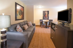 Habitación de hotel con un sofá gris, televisión, lámpara, mesa de comedor con sillas y piso alfombrado.