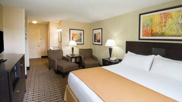 A hotel room with a large bed, two armchairs, a desk with a lamp, a telephone, and a painting on the wall.