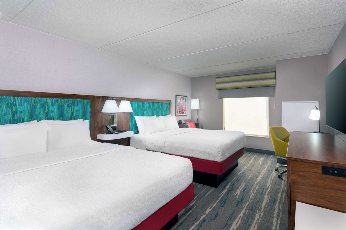 A hotel room with two beds, a desk, a chair, a TV, and a window with a view.