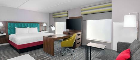 A hotel room with a bed, desk, chair, sofa, TV, and windows.