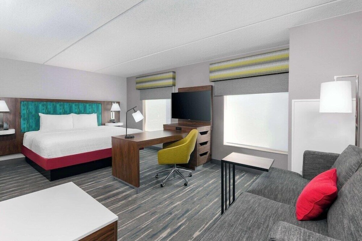 A hotel room with a bed, desk, chair, TV, sofa, and large windows.