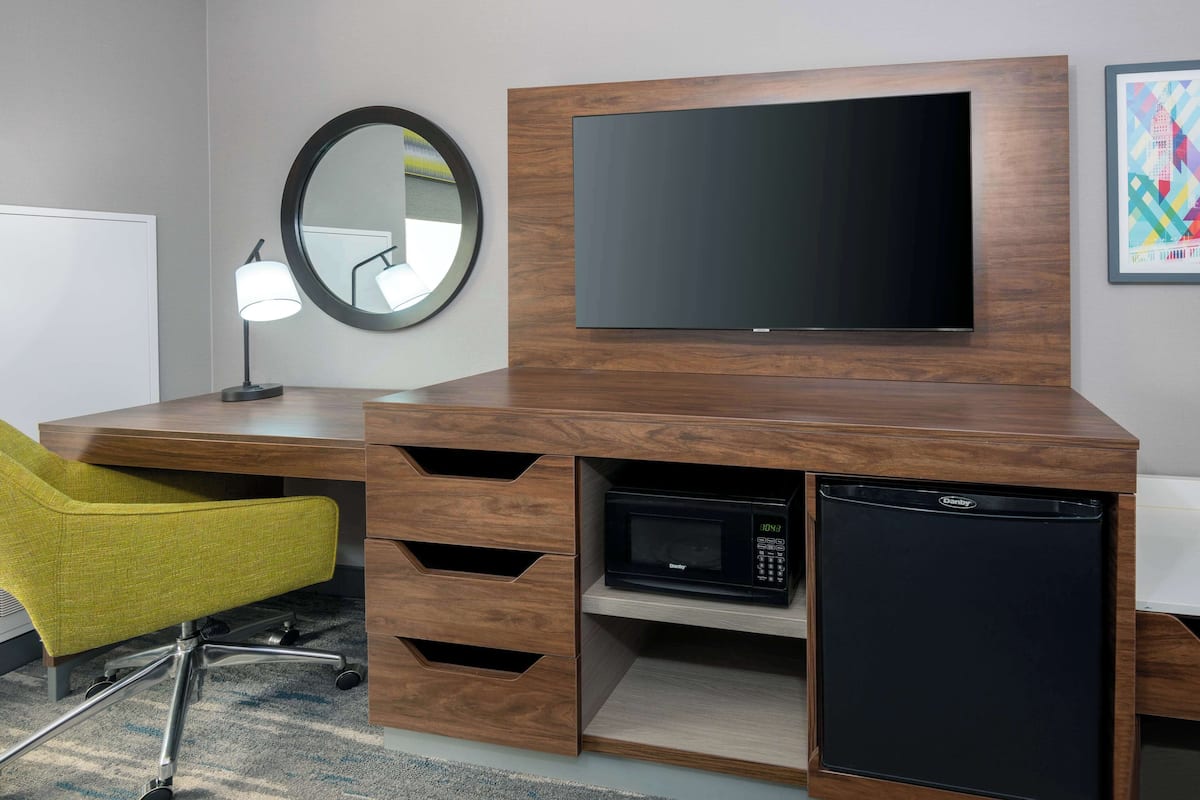 A modern hotel room with a wooden desk, a flat-screen TV, a microwave, and a compact refrigerator.