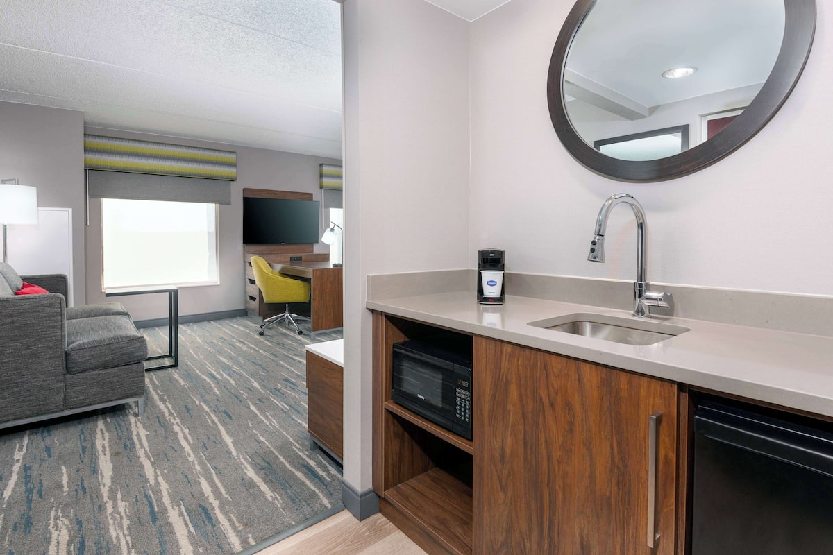 A modern hotel room with a grey sofa, a wooden cabinet with a built-in microwave, a sink, and a mirror.