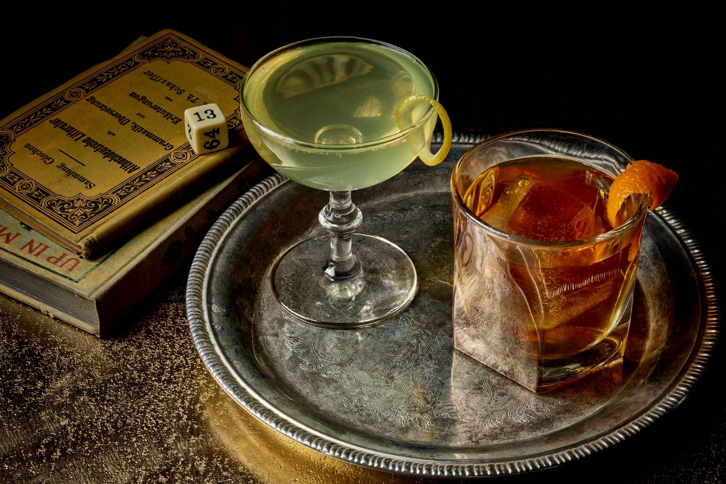 A tray with a cocktail glass and a glass of tea, a book, and a dice.