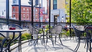An outdoor cafe with metal chairs and tables, a view of a red brick building, and a tree in the background.