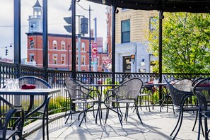 An outdoor cafe with metal chairs and tables, a view of a red brick building, and a tree in the background.