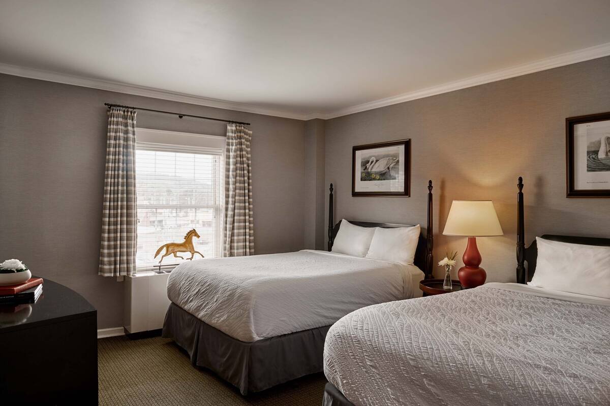 A hotel room with two beds, a window with curtains, a lamp, and framed pictures on the wall.