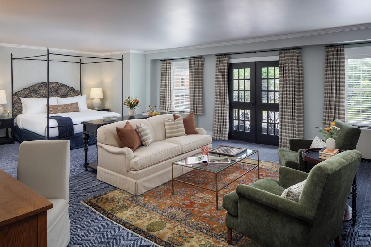A hotel room with a four-poster bed, a sofa, a green armchair, a coffee table, and a view of the outdoors through large windows with curtains.
