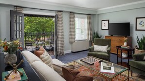 A living room with a sofa, armchairs, a coffee table, and a television. There is a balcony with a view of trees.