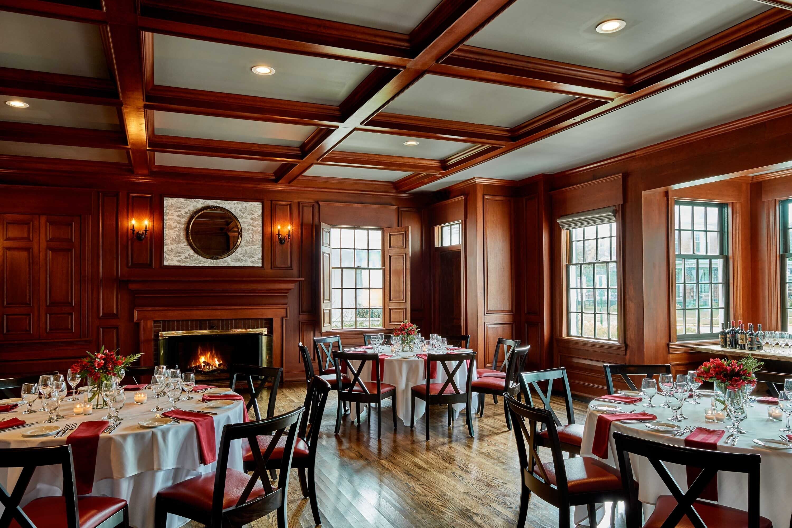 A dining area with wooden paneling, a fireplace, and large windows. Tables are set with white tablecloths and red napkins.
