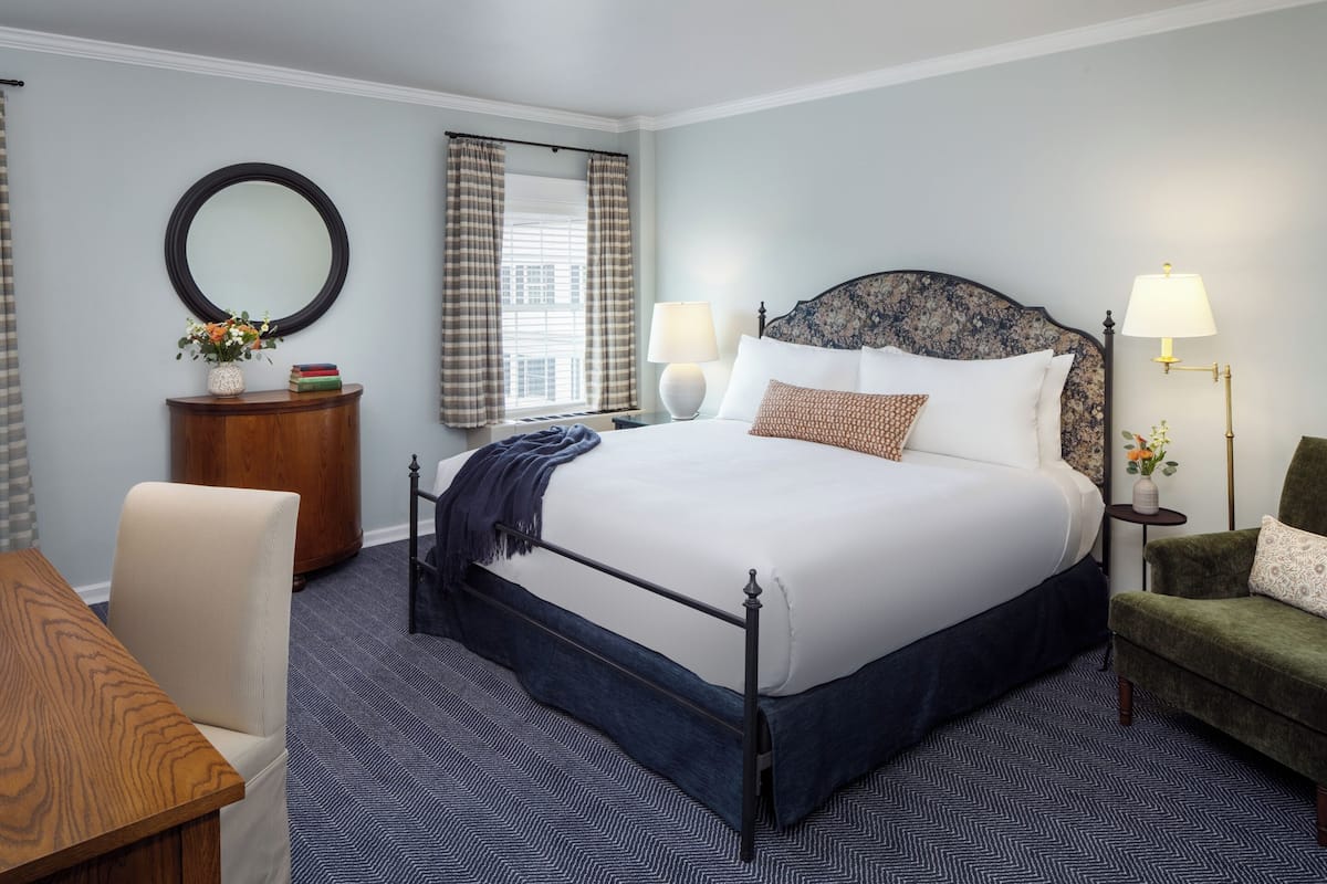 A neatly arranged hotel room with a large bed, two bedside lamps, a round mirror, a small side table, and a chair.