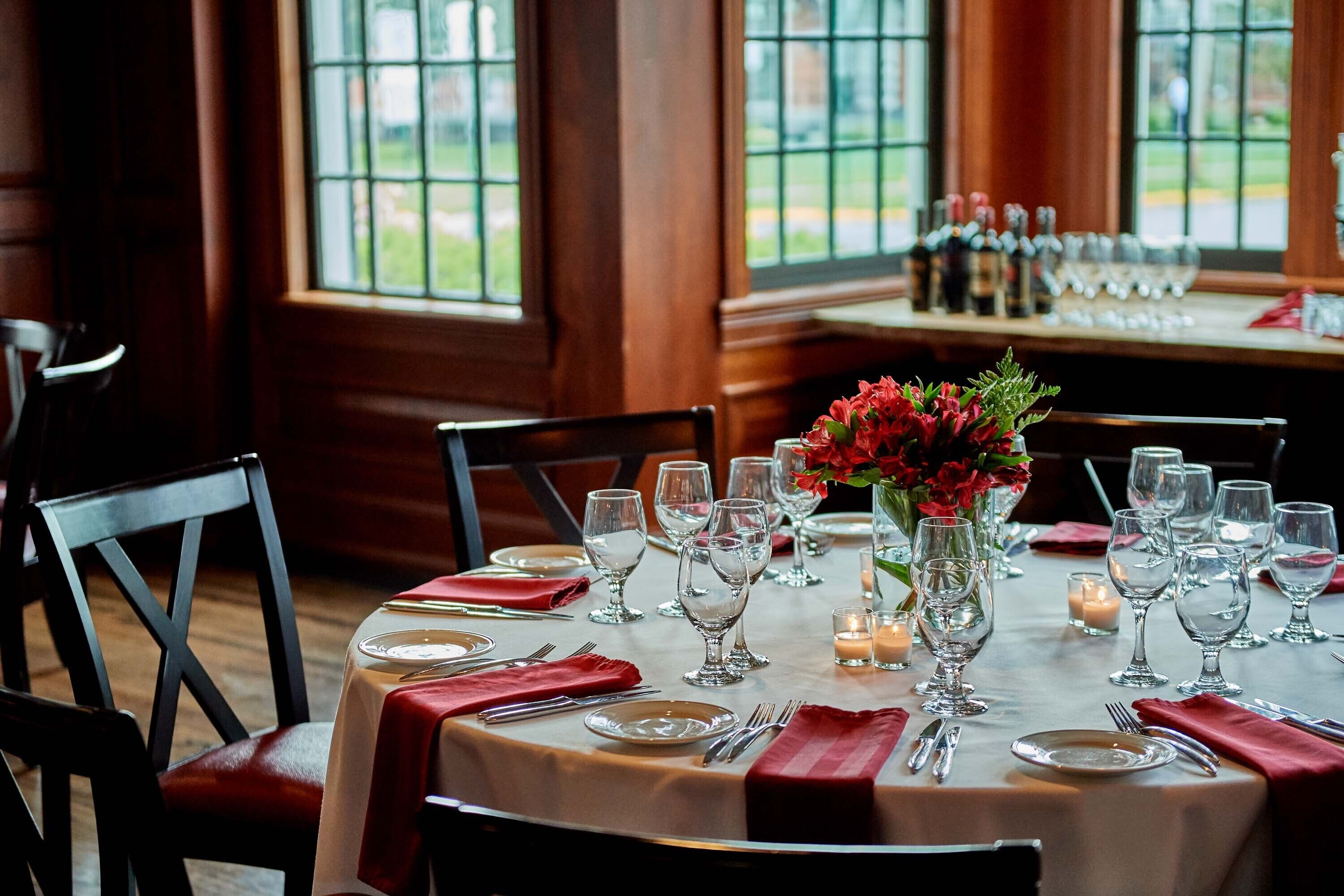 A dining table set with red napkins, clear glassware, and a vase of red flowers.
