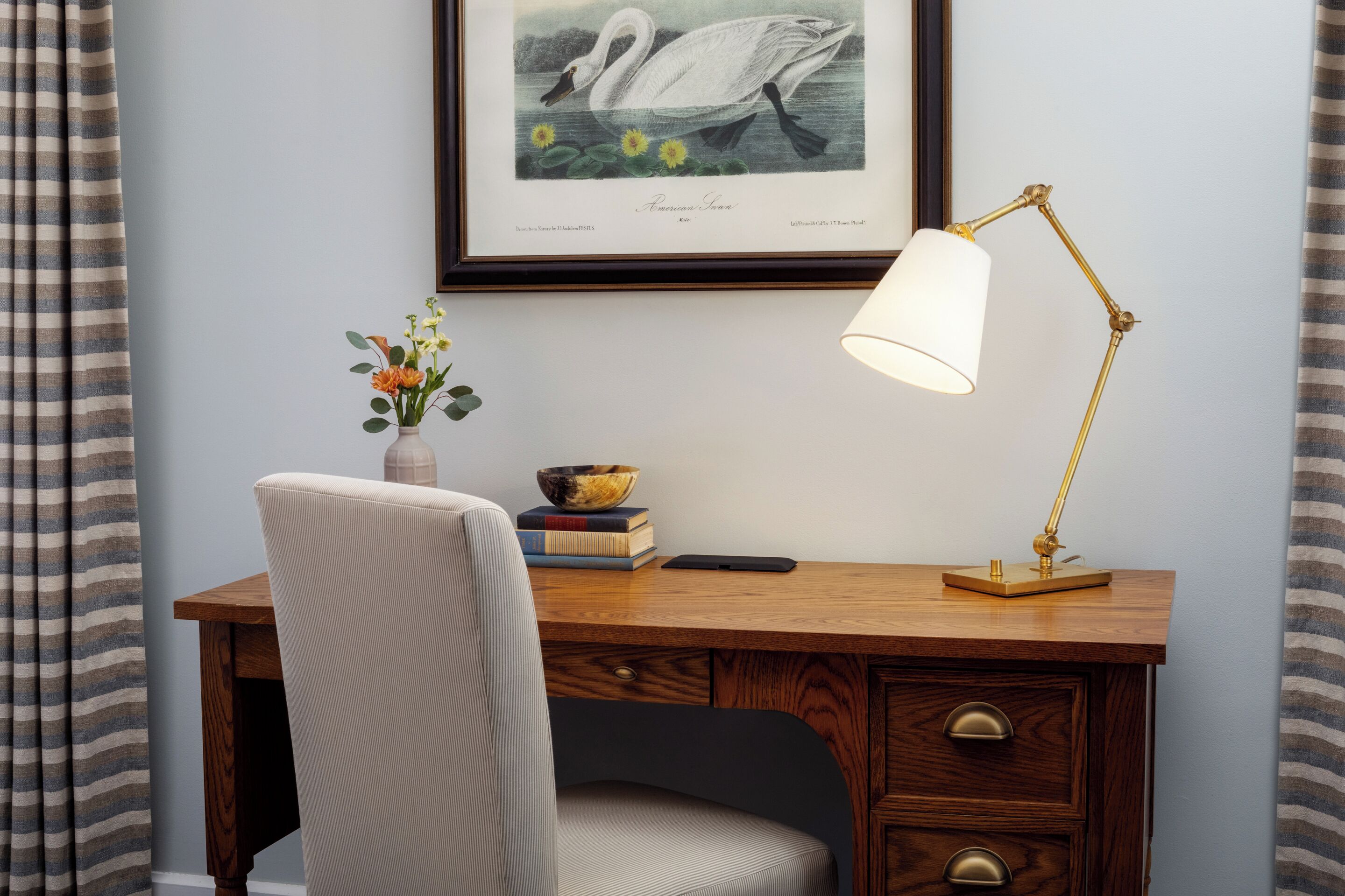 A wooden desk with a chair, a framed swan artwork, a lamp, a vase with flowers, and a bowl on the desk.