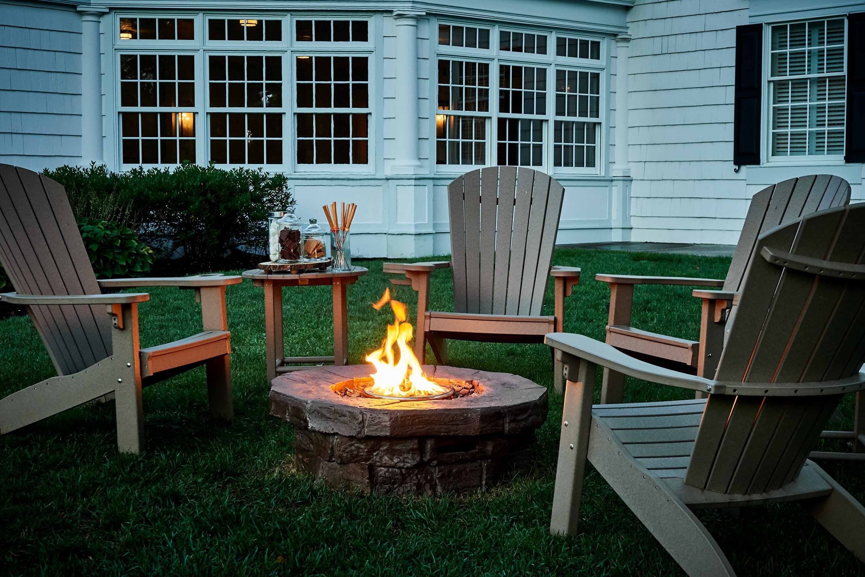 A stone fire pit with chairs around it, set in a grassy area with a white building in the background.