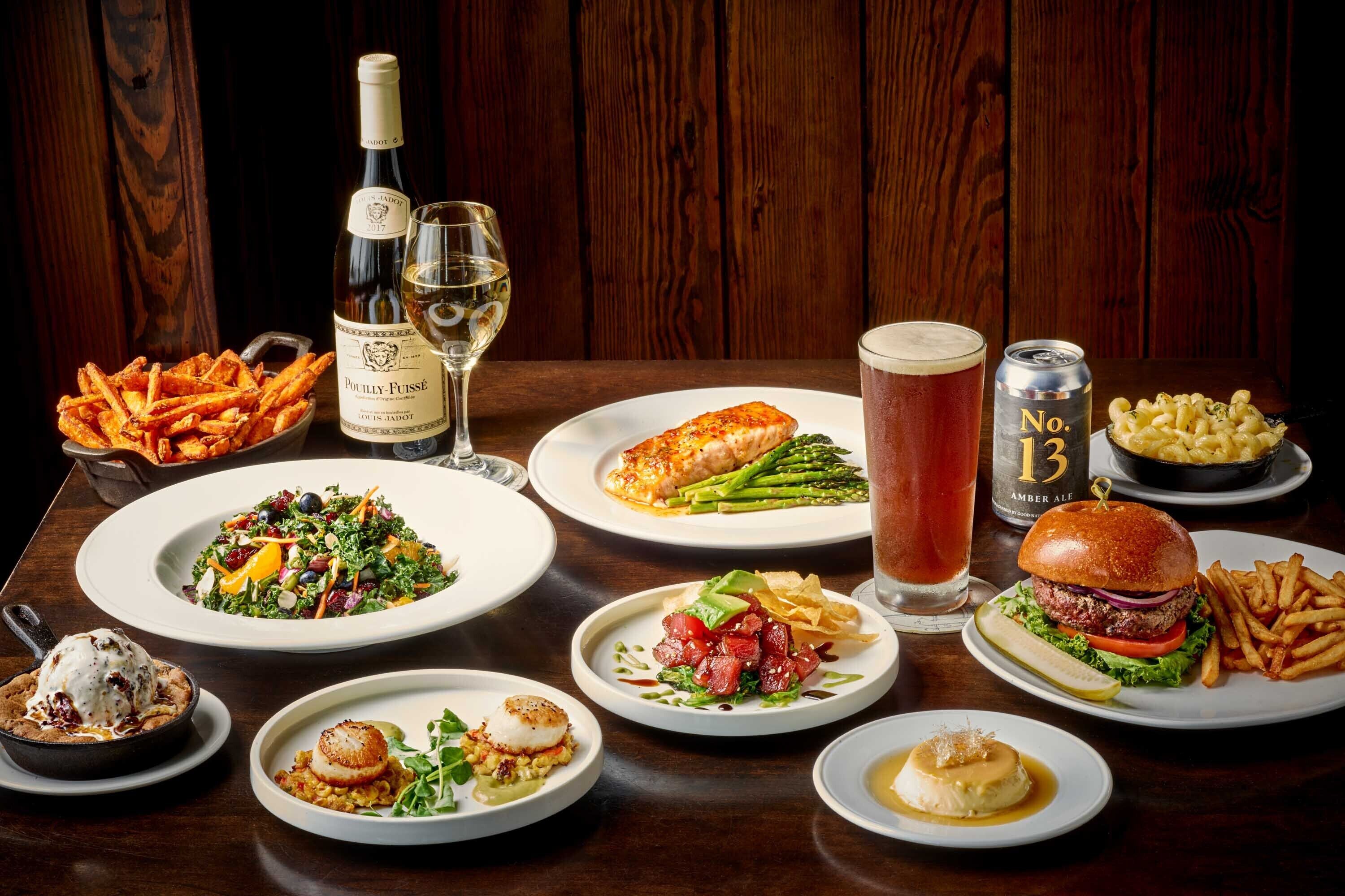 Variedad de platos que incluyen carne, pasta y mariscos, dispuestas sobre una mesa de madera con una botella de vino y una lata de cerveza.