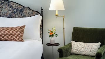 A neatly made bed with white and patterned pillows, a green armchair, a small table with a vase of flowers, and a floor lamp.